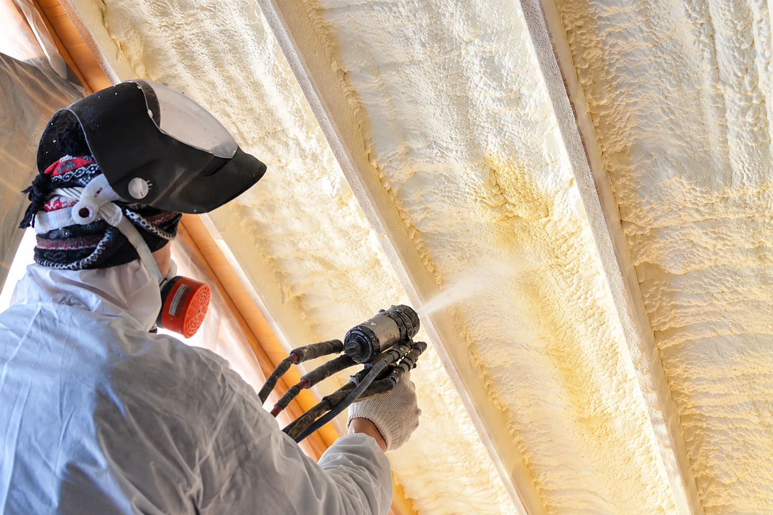 A worker in protective gear sprays foam insulation onto the underside of a roof.