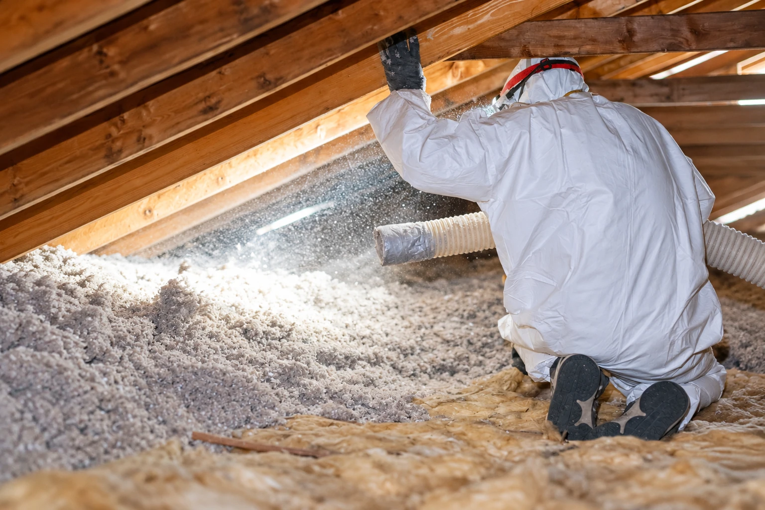 A worker in protective coveralls sprays blown-in insulation across an attic floor through a hose.