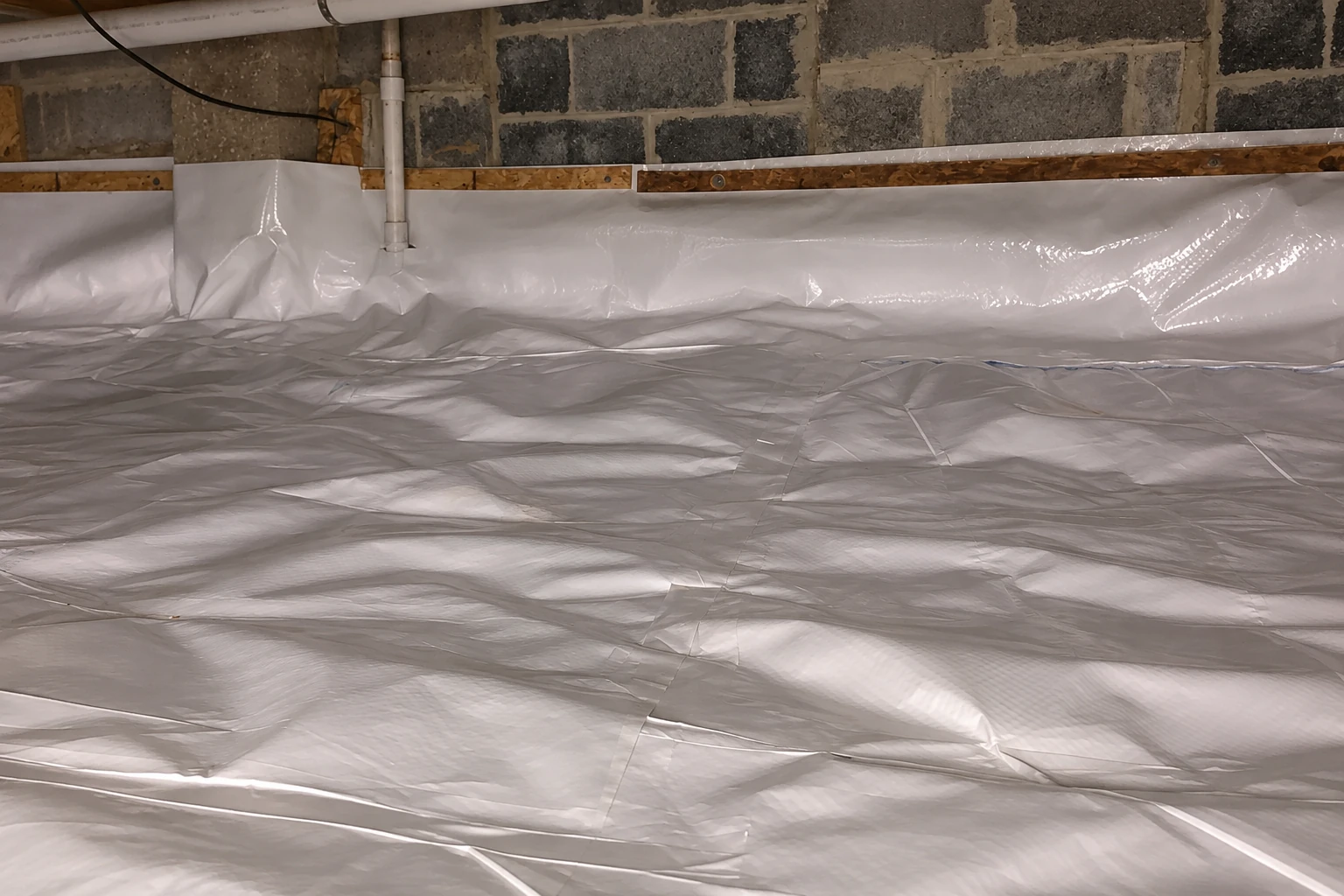 Crawl space floor covered with overlapping gray vapor barrier, with white sheeting lining a stone foundation wall.