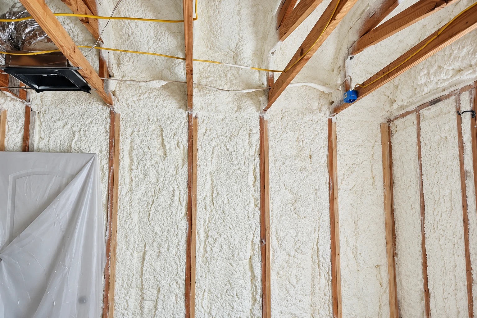 Open-cell spray foam insulation coats the walls and roof between exposed wooden framing in an unfinished room.