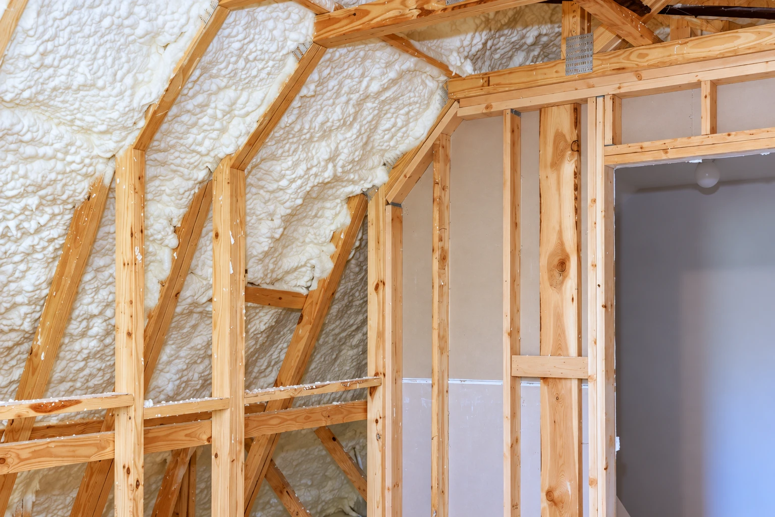 Spray foam insulation fills the sloped roof framing between exposed wooden rafters in an unfinished room.